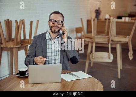 Joyful man using notebook and talking on cellphone in cafe Stock Photo