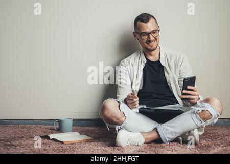 Smiling Caucasian man sitting on the floor at home looking at cellphone screen watch an online conferice making notes. Internet technology Stock Photo