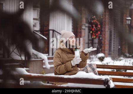 Cheerfully brunette woman drinking from flask outdoors in the city in winter. Warming up, enjoying moment, having brake over snow covered trees Stock Photo