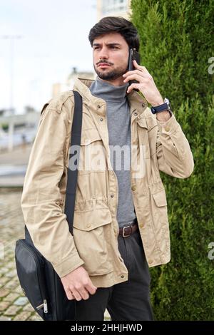 Concerned bearded male in outerwear standing on city street and having serious conversation on cellphone Stock Photo