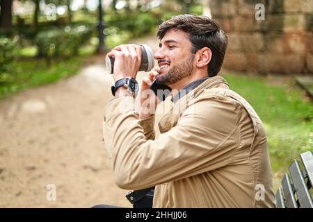 Side view of positive bearded male student sitting with laptop bag and drinking coffee while talking on cellphone on bench in park Stock Photo