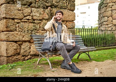 Full body of positive bearded male student sitting with laptop bag and drinking coffee while talking on cellphone on bench in park Stock Photo