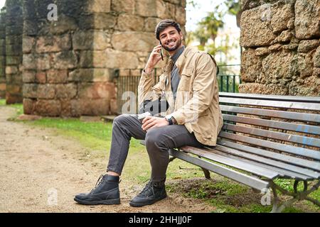 Full body of positive bearded male student sitting with laptop bag and drinking coffee while talking on cellphone on bench in park Stock Photo
