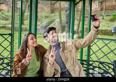 Happy friends in outerwear sitting on bench in alcove and showing v sign while taking self portrait on cellphone in park Stock Photo