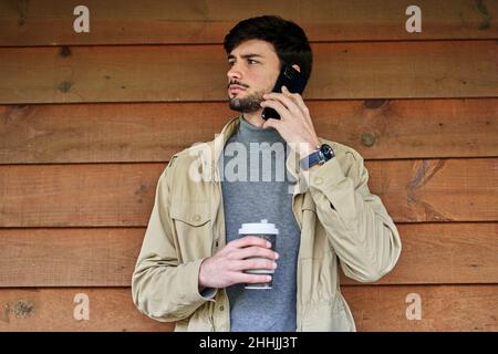 Thoughtful bearded male in casual clothes standing near wooden wall with cup of hot beverage and talking on mobile phone Stock Photo
