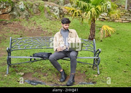 Full body of serious bearded male student typing on netbook while preparing for exams in green park Stock Photo