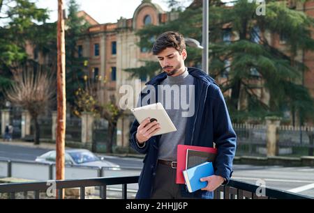 Bearded male student holding notebooks and folder for studies and checking tablet on city street Stock Photo