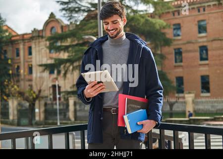 Happy bearded male student holding notebooks and folder for studies and checking tablet on city street Stock Photo