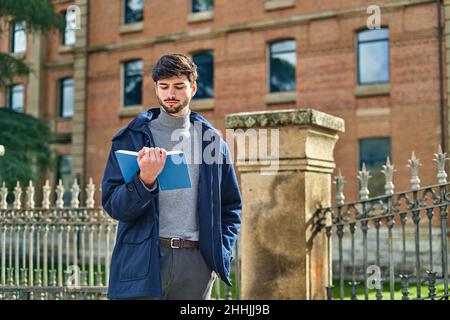 Bearded male student in outerwear standing with hand in pocket and reading textbook for homework in university campus Stock Photo