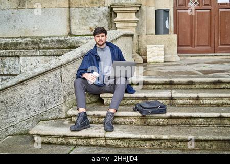 Male student sitting on stone staircase and browsing netbook while preparing for university task and having coffee Stock Photo