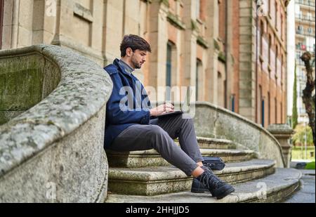 Side view male student sitting on stone staircase and browsing netbook while preparing for university task Stock Photo