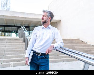 Confident male entrepreneur with modern netbook in hand leaning on railing while standing on staircase near modern office building in city Stock Photo