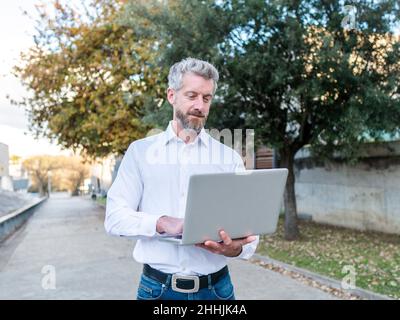 Focused bearded male entrepreneur in white shirt typing on modern netbook while standing on street during online work in city Stock Photo