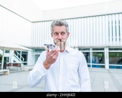 Bearded male traveler in white shirt recording audio message on smartphone standing near luggage on street with building in city Stock Photo