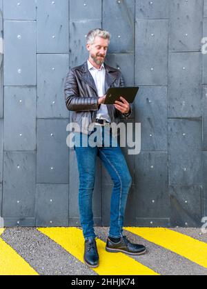 Full body of content male surfing internet on modern black tablet while standing near gray wall on street of city Stock Photo