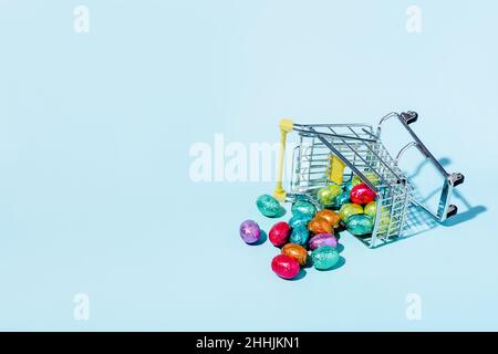 An overturned shopping cart on the ground and spilling colourful Easter eggs on a blue background. Stock Photo