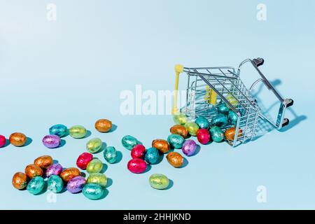 An overturned shopping cart on the ground and spilling colourful Easter eggs on a blue background. Stock Photo