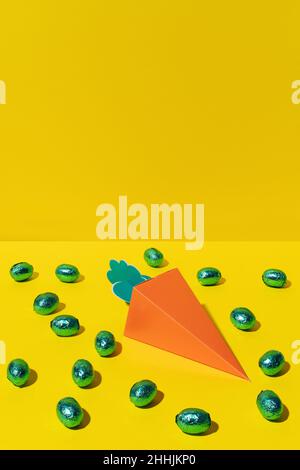Cardboard carrot on a yellow background in the daylight surrounded by green chocolate foil eggs at easter Stock Photo