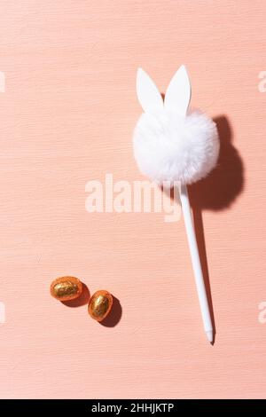 A white pen with a pompon on an orange background and chocolate eggs. The pen looks like a rabbit Stock Photo