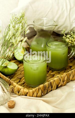 High angle glasses and carafe of fresh green smoothie with ice cubes served on wicker tray with sliced apple and gentle flowers on bed Stock Photo