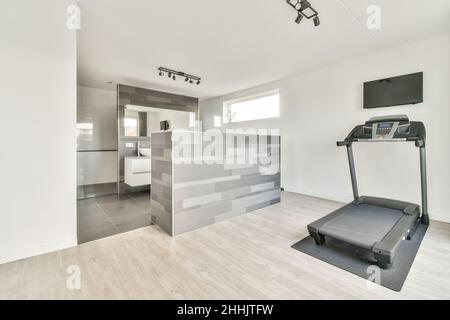 Modern treadmill in corridor with TV set hanging on white wall near entrance to tilled bathroom in big house Stock Photo