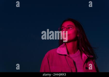 Attentive teen female with long brown hair in eyeglasses admiring dark night in pink illumination Stock Photo
