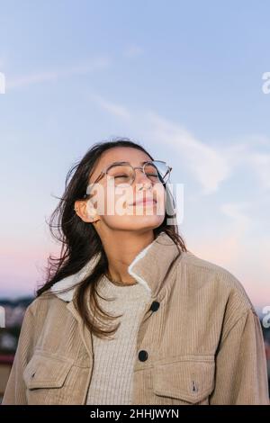 Happy teen female in eyeglasses smiling widely with eyes closed against sundown sky in evening Stock Photo