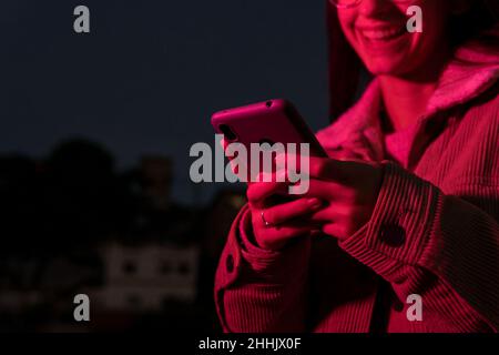 Cropped unrecognizable happy teen female in outerwear and eyeglasses laughing brightly while reading merry message on smartphone on dark street illumi Stock Photo