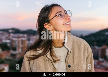 Happy teen female in eyeglasses smiling widely while looking up against sundown sky in evening Stock Photo