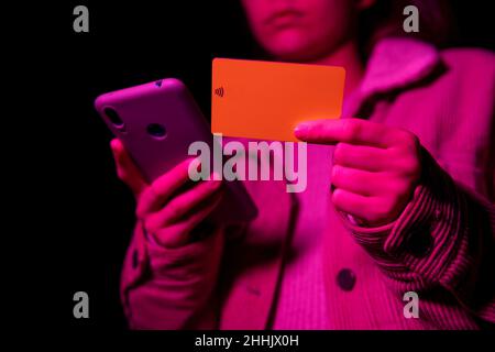 Low angle of crop teenage female paying for online purchases with credit card via cellphone at dark night with pink illumination Stock Photo