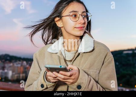 Crop teenage female in eyeglasses browsing cellphone while leaning on fence on rooftop in windy evening at sunset and looking away Stock Photo