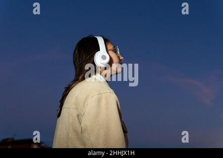 Unrecognizable side view of teen female in outerwear and eyeglasses keeping hands on wireless headphones and enjoying good music outdoors at sundown Stock Photo
