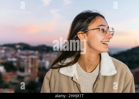 Happy teen female in eyeglasses smiling widely while looking up against sundown sky in evening Stock Photo