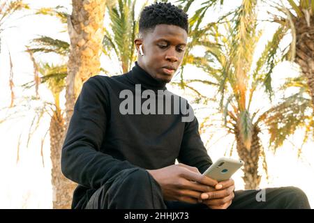 Young black guy sitting on bench in summer park and browsing cellphone and earbuds in sunlight Stock Photo