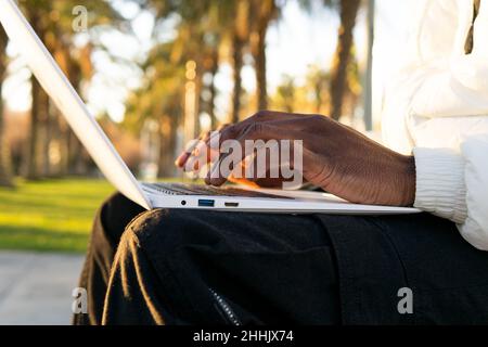 Cropped unrecognizable African American freelancer sitting on bench in park and browsing laptop while working remotely on sunny day Stock Photo