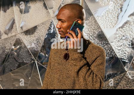 Unemotional bearded African American male having phone conversation while standing near wall on sunny street in city Stock Photo