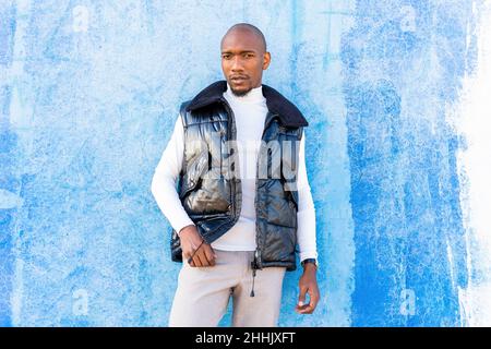 Handsome bald African American male in stylish wear looking at camera while standing near wall on street of city Stock Photo