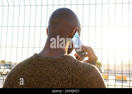 Back view of bald unrecognizable African American male having phone conversation while standing near metal fence on sunny street in city Stock Photo