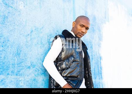 Handsome bald African American male in stylish wear looking at camera while standing near wall on street of city Stock Photo