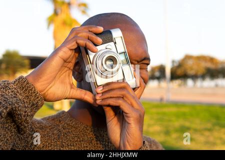 Focused bald African American male photographer taking picture on photo camera while standing on grassy lawn in city with trees Stock Photo