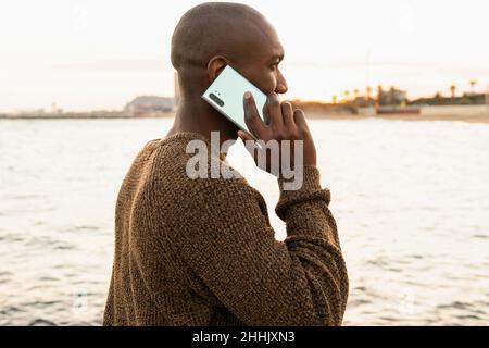 Side view of bald African American male speaking on cellphone and looking into distance while standing on coast near rippling sea Stock Photo