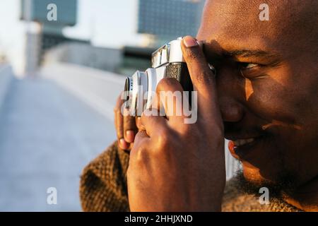 Side view of focused cheerful bald African American male photographer taking picture on photo camera while standing on grassy lawn in city with trees Stock Photo