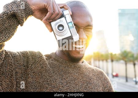 Focused cheerful bald African American male photographer taking picture on photo camera while standing on grassy lawn in city with trees Stock Photo