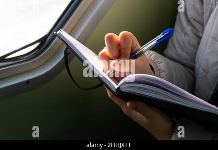 From above of crop unrecognizable female in warm jacket taking notes in planner sitting near window in boat Stock Photo