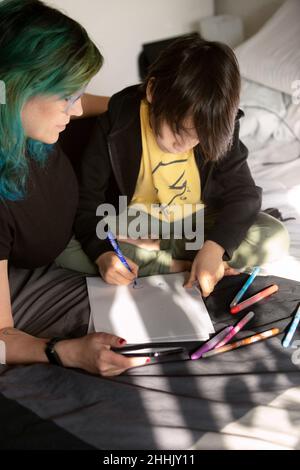 Caring mother sitting near focused talented son drawing on paper with felt pen on comfortable bed in light bedroom at home Stock Photo