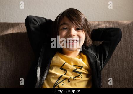 Cheerful ethnic boy in casual clothes looking at camera with hands behind head while sitting on sofa Stock Photo