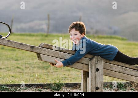 Happy boy looking at camera while lying on wooden seesaw while playing on playground with grassy lawn during mountain ridge Stock Photo