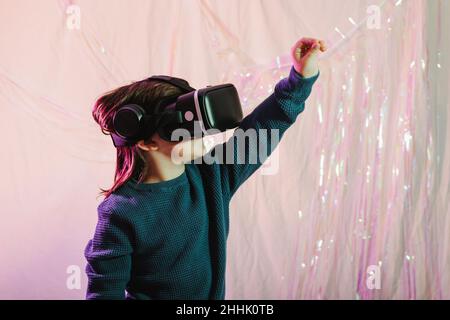 Side view of excited girl in VR headset touching invisible object while experiencing virtual world on white shiny background Stock Photo