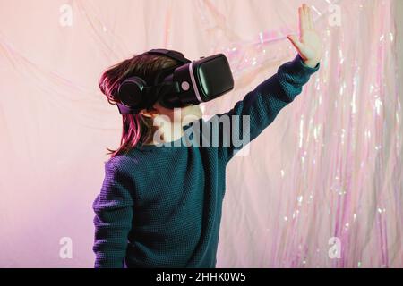 Side view of excited girl in VR headset touching invisible object while experiencing virtual world on white shiny background Stock Photo