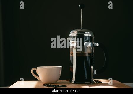 White ceramic cup with French press coffee pot with fresh roasted beans served on wooden table against black background Stock Photo